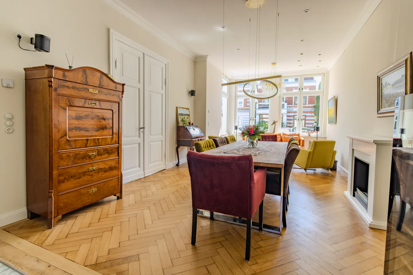 Dining room with herringbone wood floors, a long table, colorful upholstered chairs, and a large antique wooden cabinet.