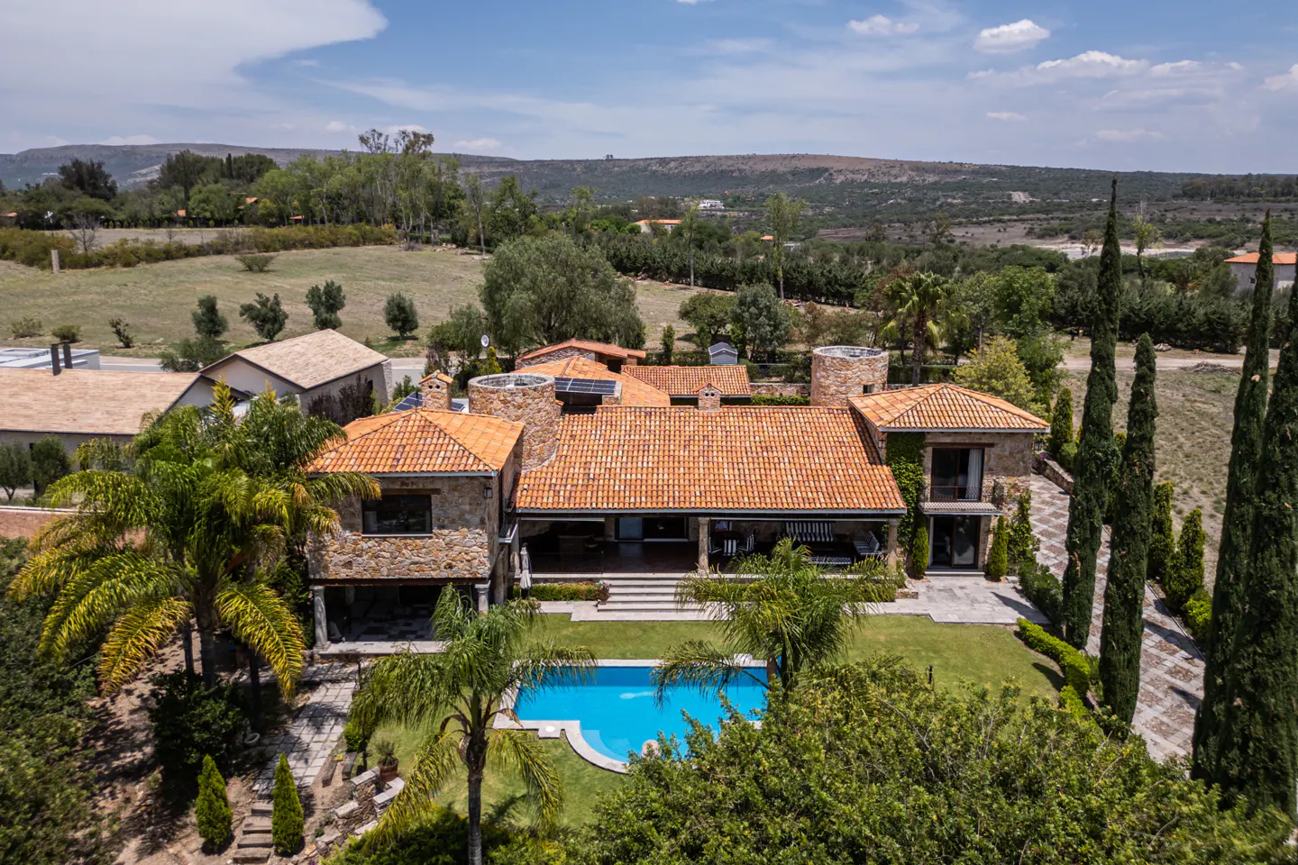 Aerial view of a stone house with a red tile roof, a blue pool, and green trees against a hilly background.