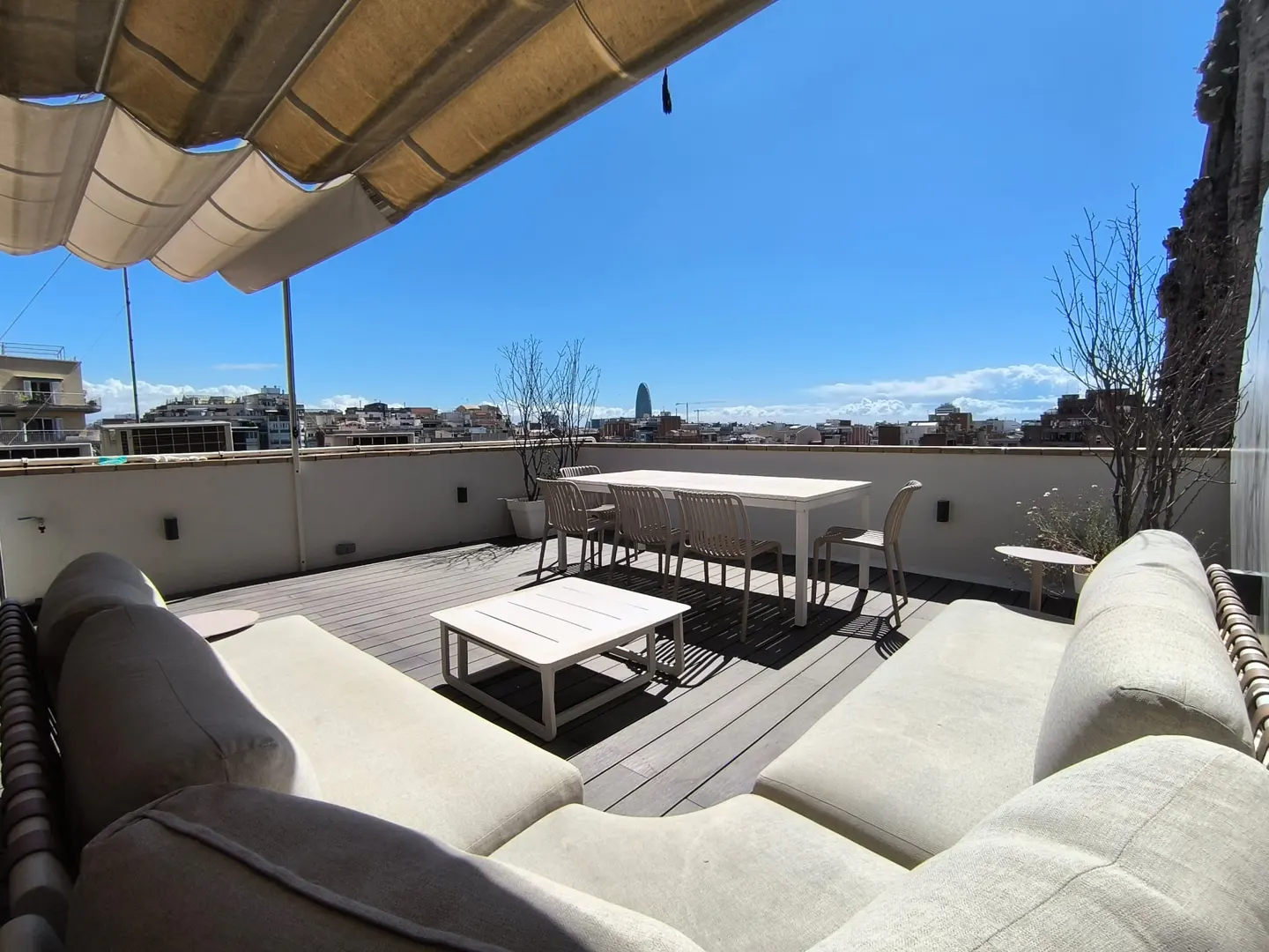 Rooftop patio with white furniture, including a large sectional sofa, dining table, and coffee table, with a city view under a bright blue sky.