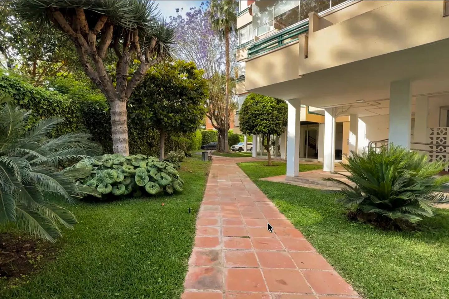 Landscaped walkway with red brick pavers leading to a light-colored apartment building with green balconies and lush green foliage.