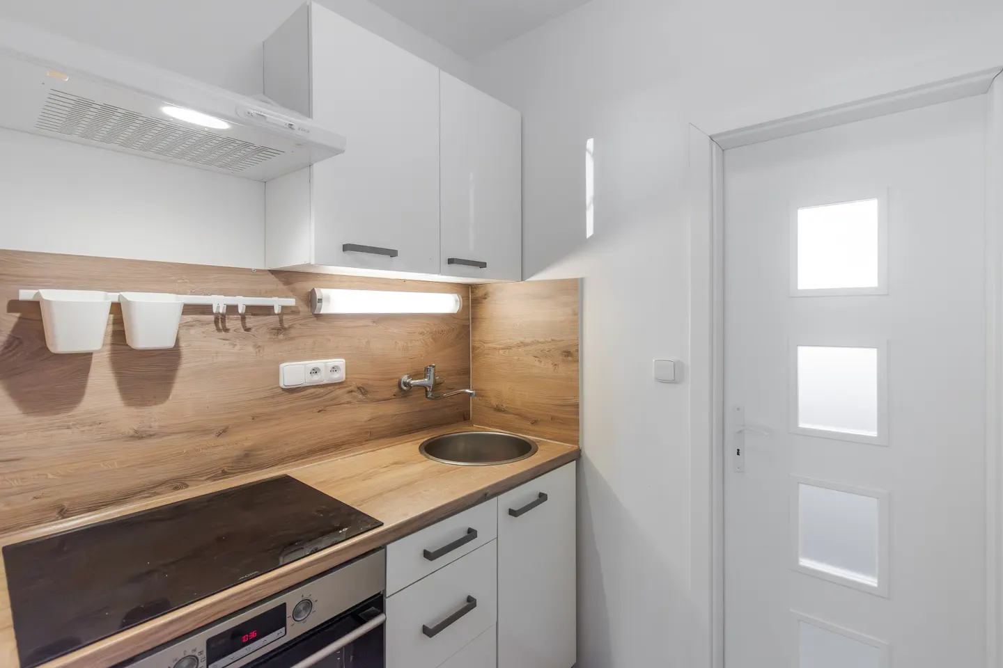 A compact kitchen with white cabinets, wood-look countertops, a stainless steel sink, and a black stovetop. A white door is on the right.