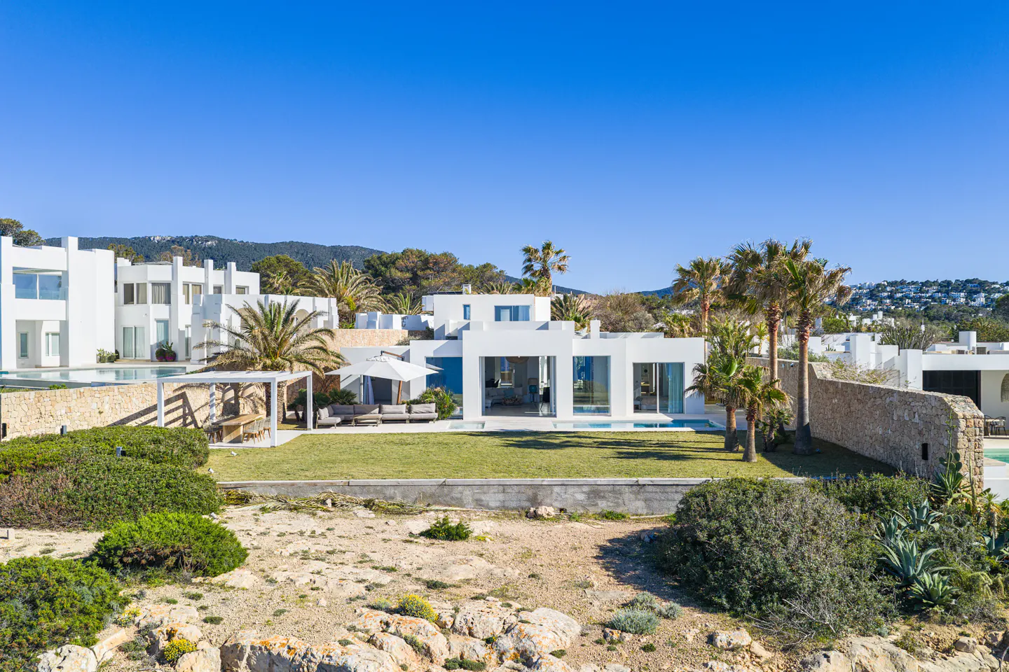 Modern white villa with palm trees and a green lawn under a clear blue sky. Stone walls and lush greenery surround the property.