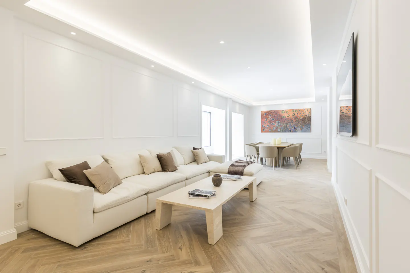 Bright living room with white walls, herringbone wood floors, and a cream sectional sofa with brown pillows. A dining table is visible in the background.