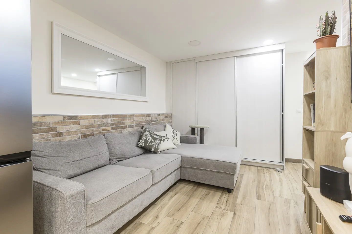 Modern living room with a grey sectional sofa, brick-style accent wall, large mirror, and light wood flooring.