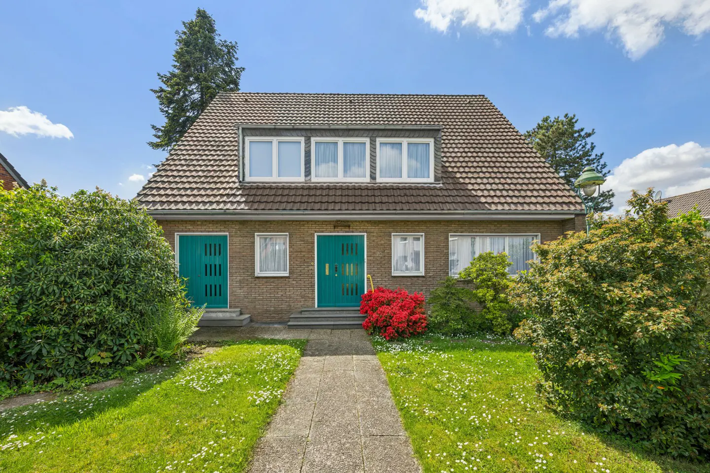 Two-story brick house with a brown tiled roof, green doors, and a stone walkway leading to the entrance. The lawn is green with white flowers.