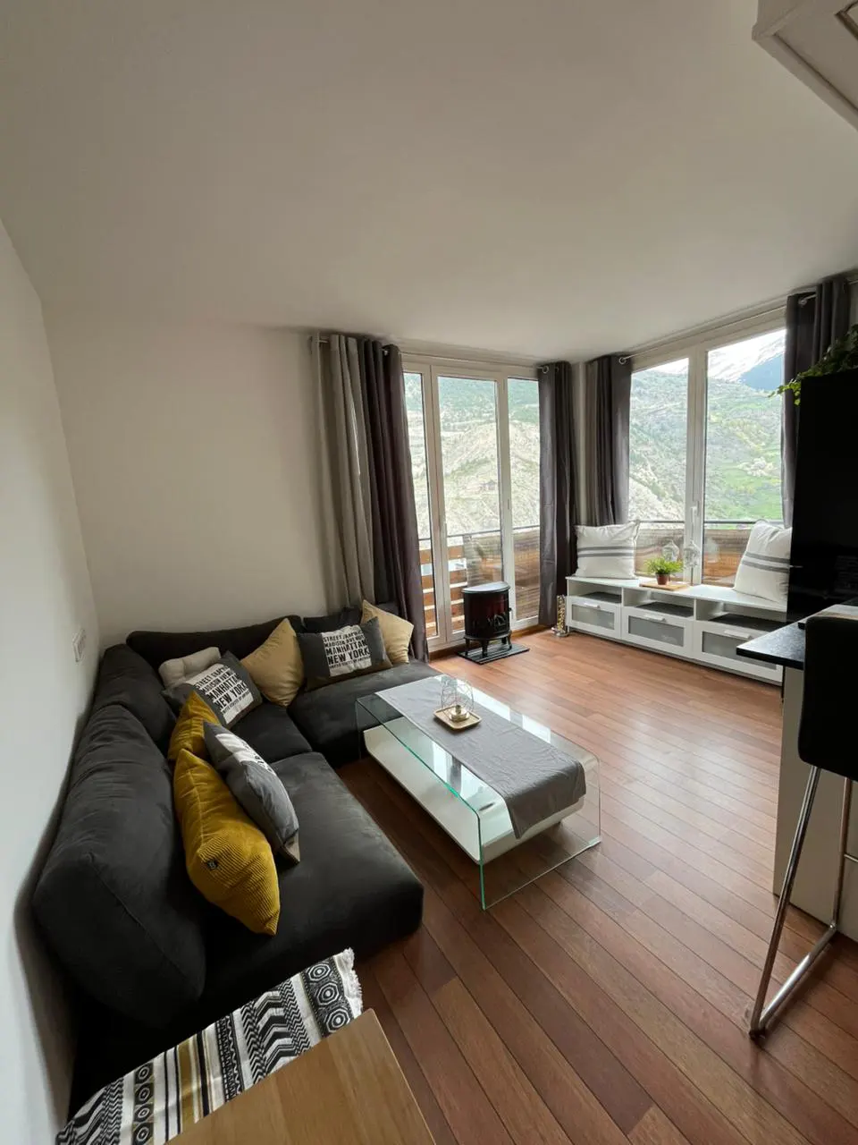 Living room with a gray sectional sofa, glass coffee table, and wood floors. Large windows offer mountain views. Gray curtains frame the windows.