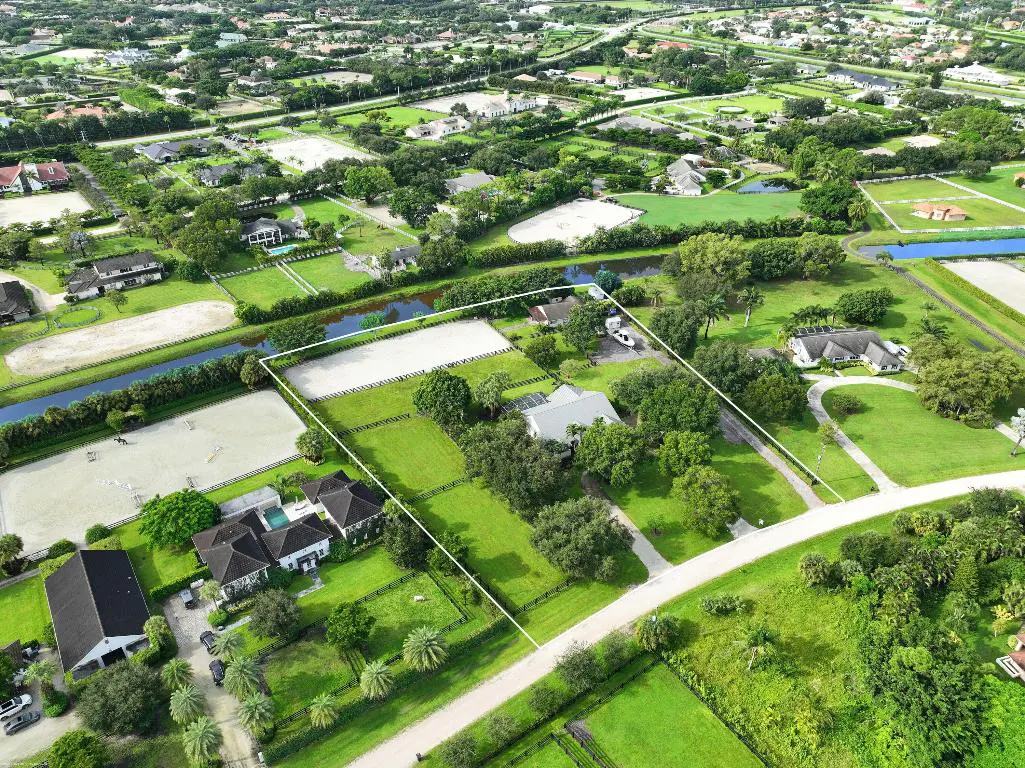 Aerial view of a large, green property with a white-lined border, featuring houses, trees, and horse riding arenas.