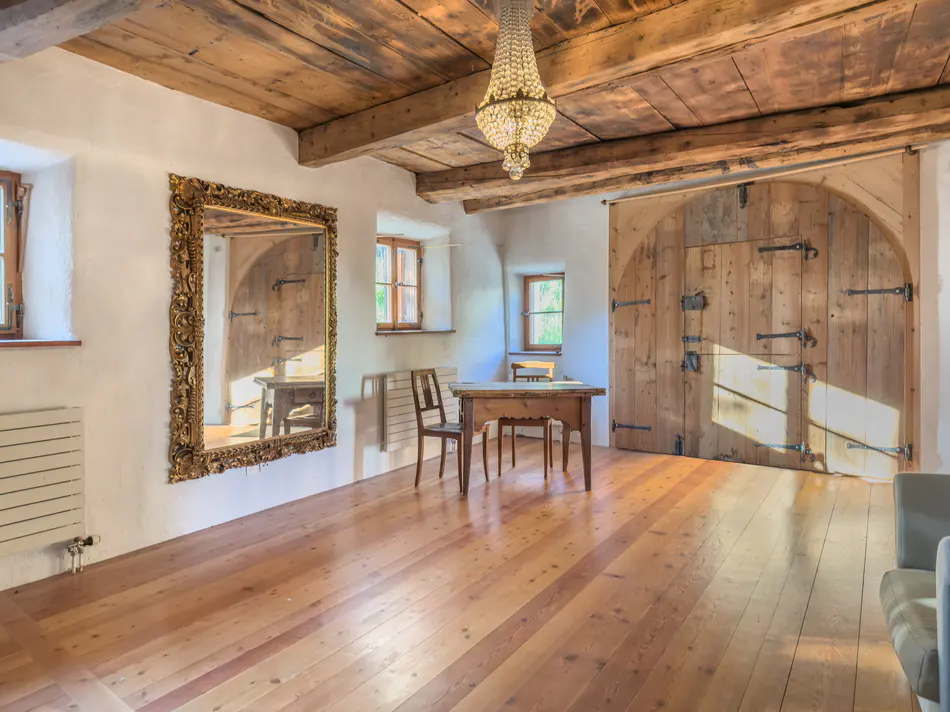 Bright room with wood floors, white walls, and wood ceiling. A large gold mirror, chandelier, table, and arched wood door are visible.