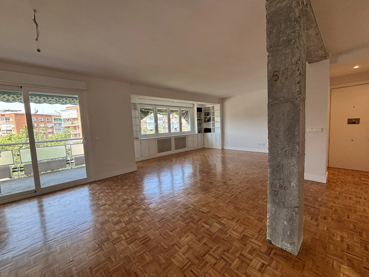 Spacious empty living room with parquet flooring, a raw concrete pillar, built-in white shelving, and a balcony door.