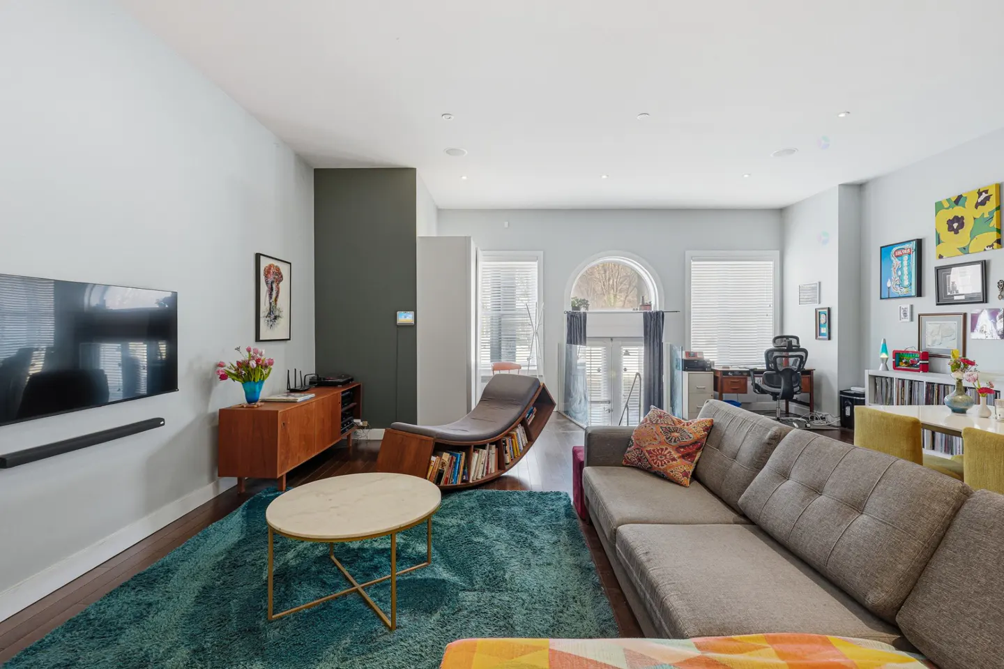 Bright living room with a gray sofa, teal rug, and a unique bookshelf chair. A TV hangs on the wall above a wooden cabinet. Artwork adorns the walls.