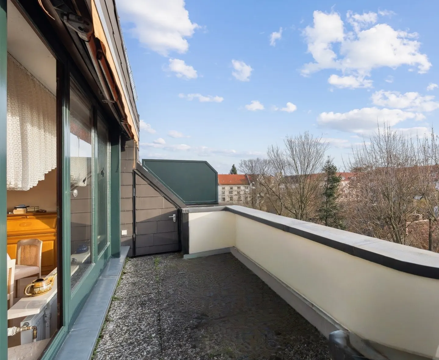 Balcony view with a white wall, dark gray floor, and green-framed windows. Buildings and trees are visible in the background under a blue sky.