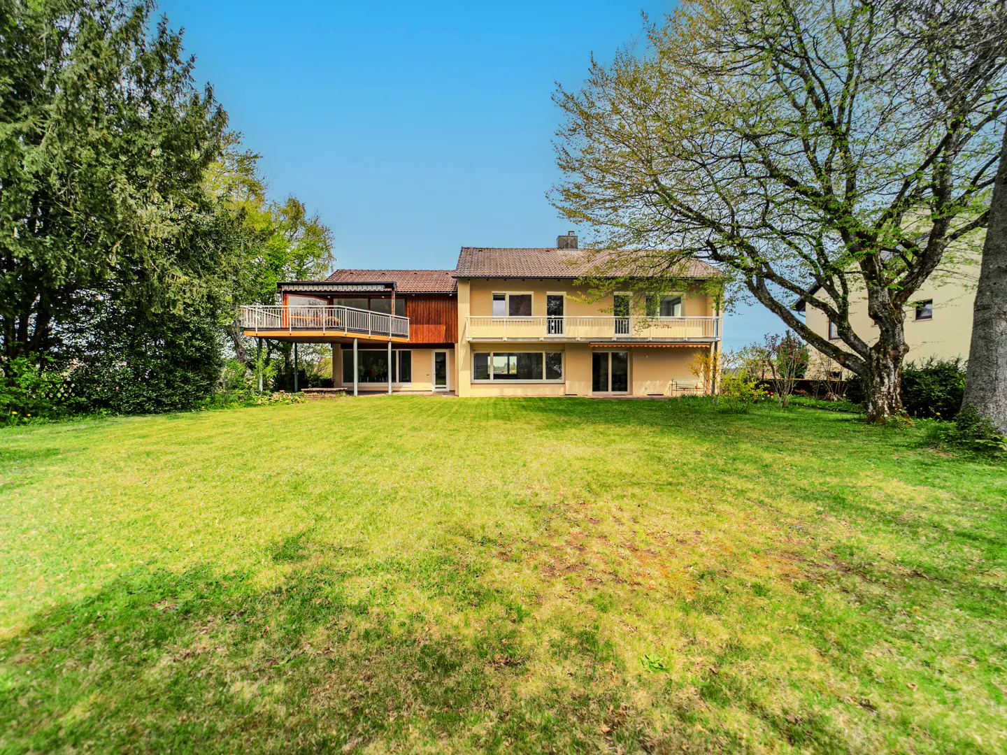 Two-story tan house with a large green lawn and trees under a blue sky. The house has a balcony and large windows.