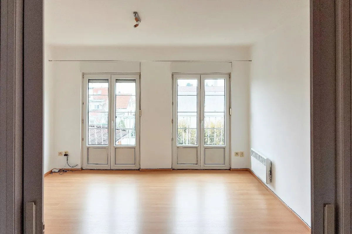 Empty room with light wood floors, white walls, and two sets of white French doors leading to balconies. A white radiator is on the right wall.