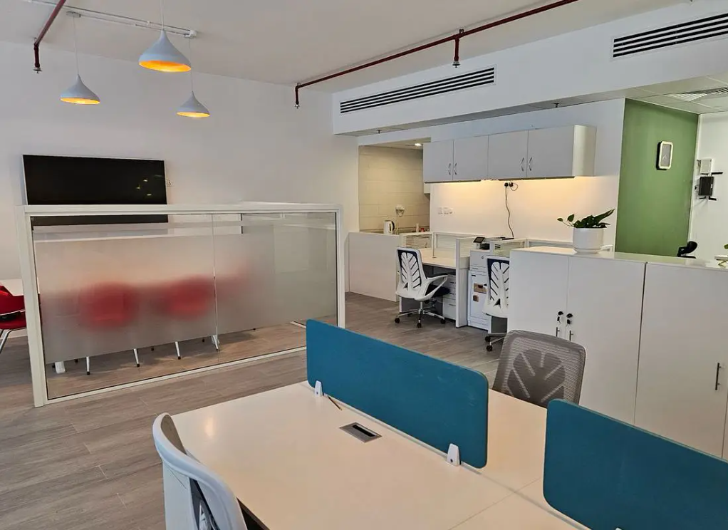 Modern office space with white desks, blue privacy dividers, ergonomic chairs, and a glass-enclosed meeting area. [AI-generated text]
