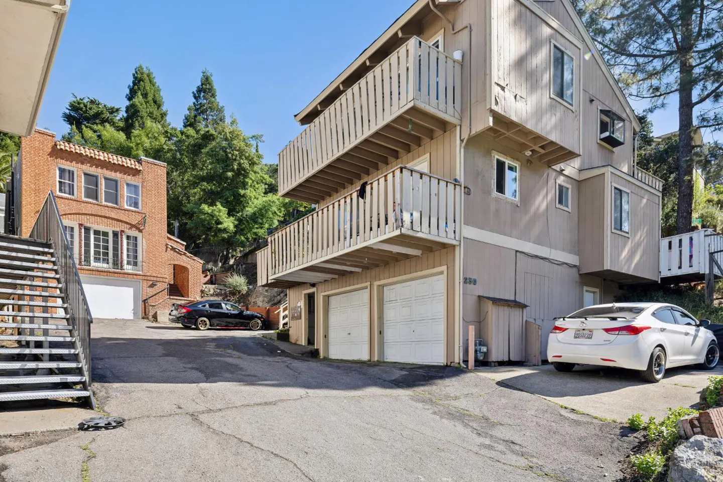 Exterior view of a tan apartment building with two balconies and garage doors, plus a brick building with stairs and parked cars.