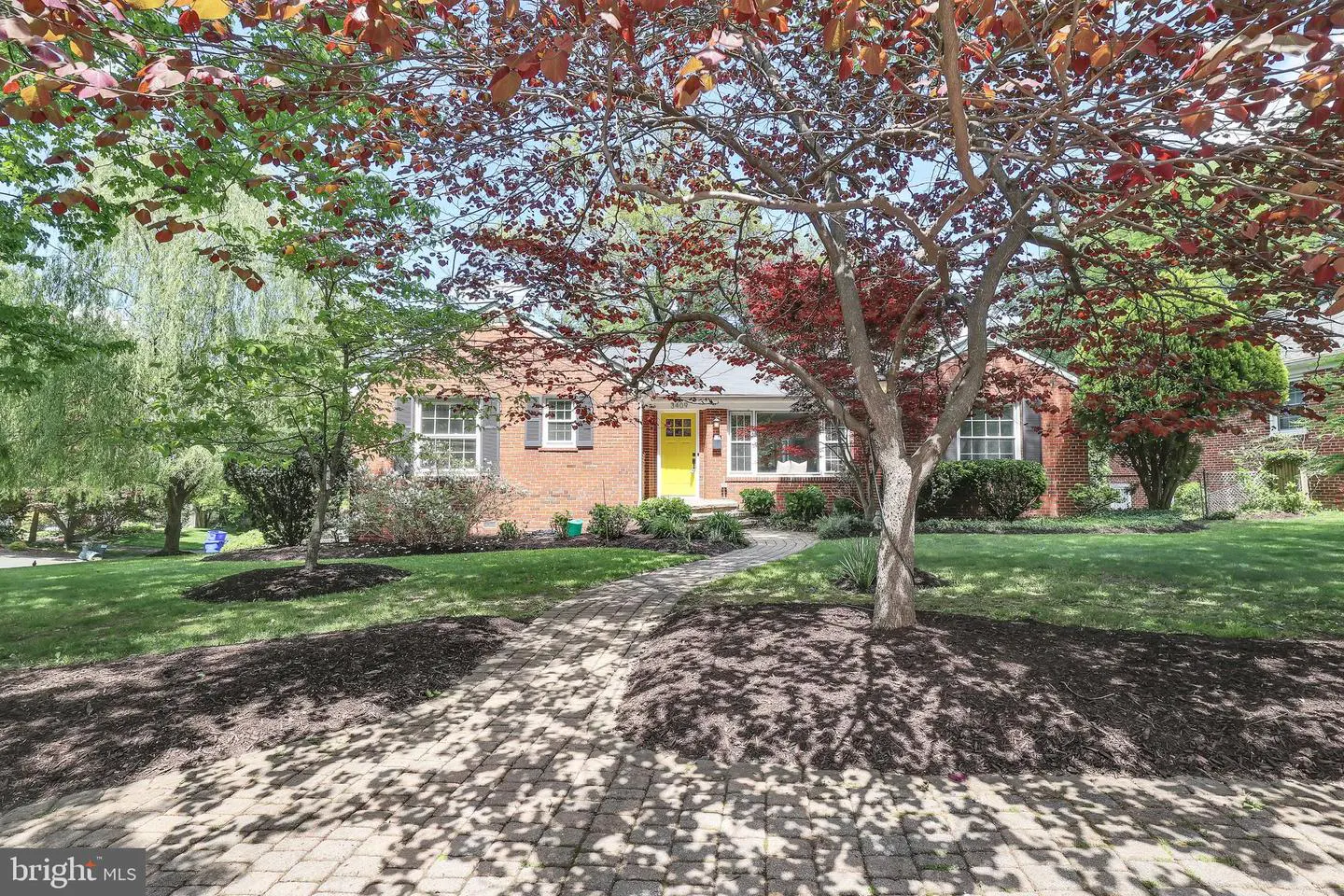 Brick house with a yellow door, viewed through trees with red and green leaves. A brick walkway leads to the entrance.
