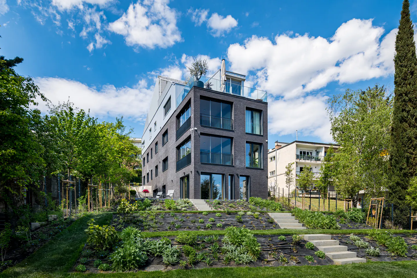 Modern black brick apartment building with a rooftop terrace and landscaped garden on a sunny day.
