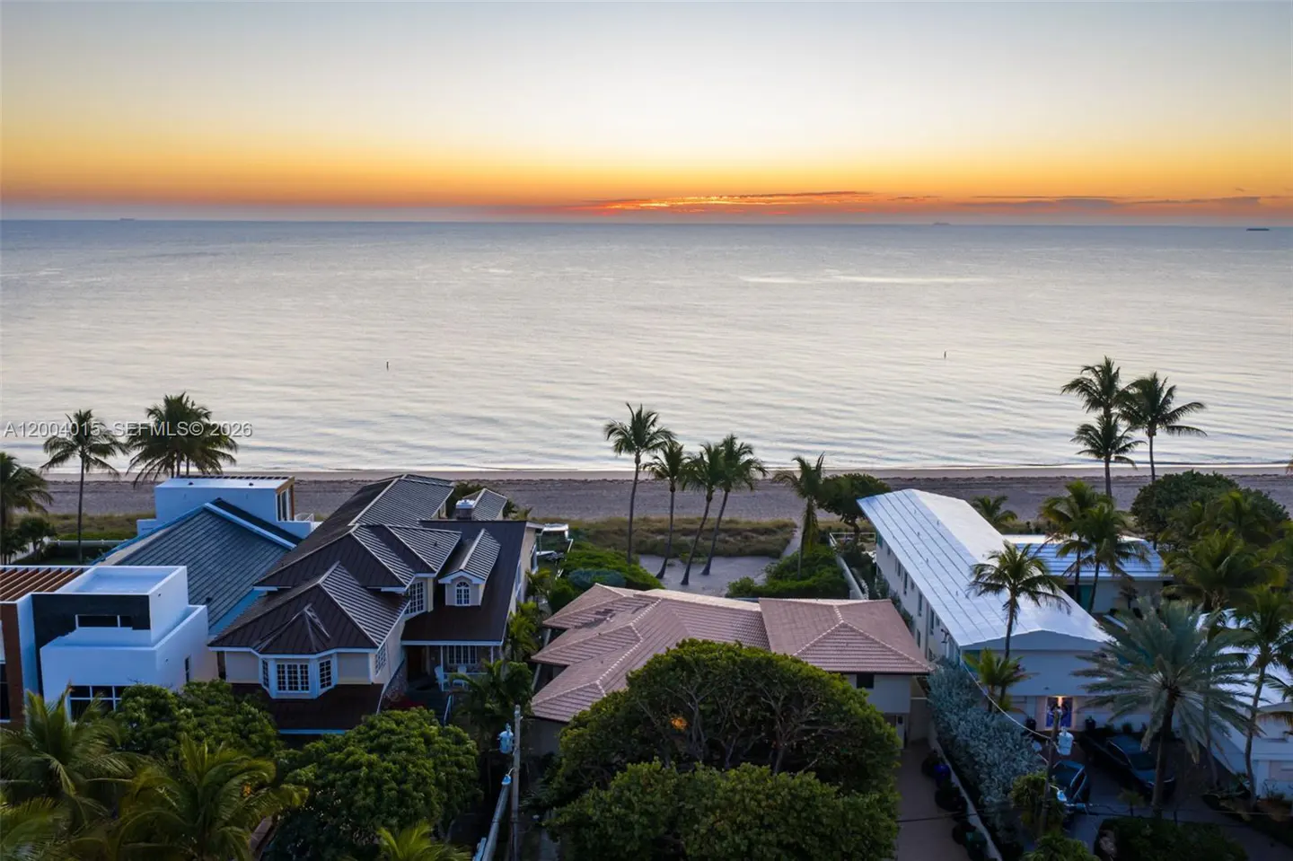 Aerial view of beachfront homes with palm trees, ocean, and a colorful sunrise sky.