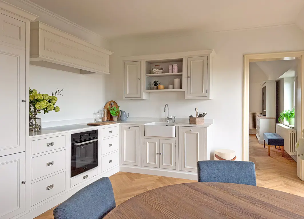 Bright kitchen with white cabinets, marble countertops, and a farmhouse sink. A round wooden table with blue chairs sits in the foreground.