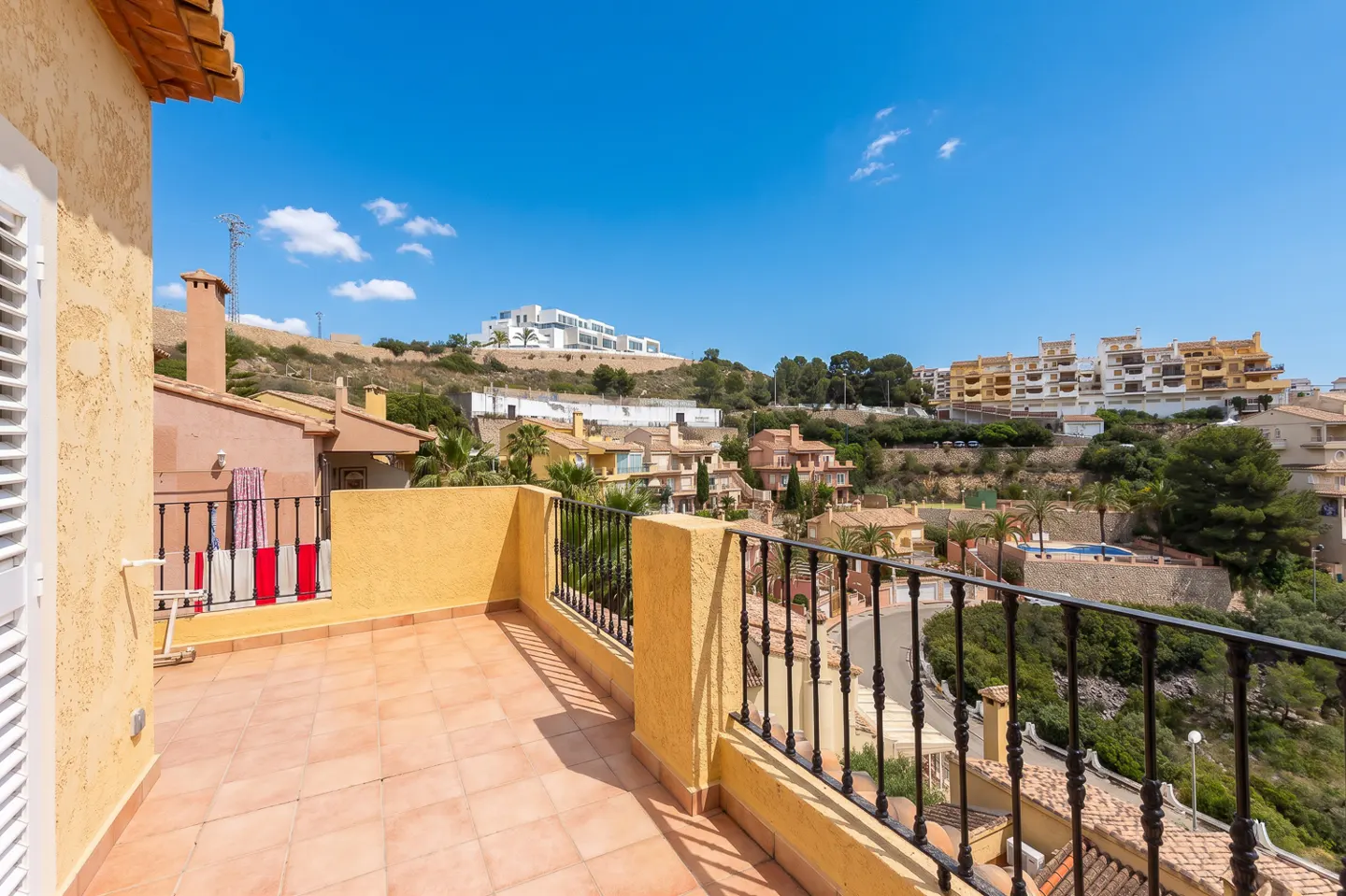 View from a sunny, tiled balcony with black railings overlooking houses on a hill under a blue sky.