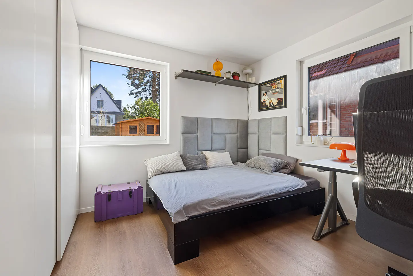 Bright bedroom with a gray bed, a purple chest, and a desk with an orange lamp. A window shows a house and shed.