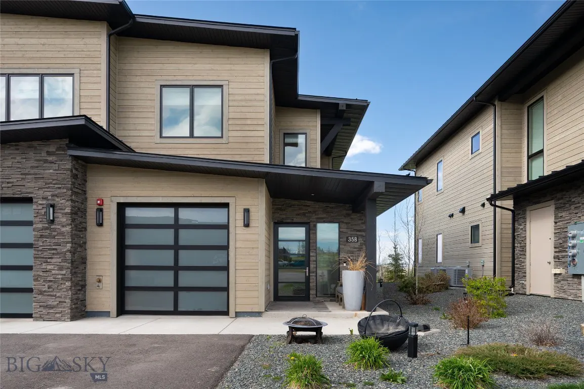 Modern townhouse exterior with beige siding, stone accents, and black trim. A glass garage door and landscaped front yard are visible.