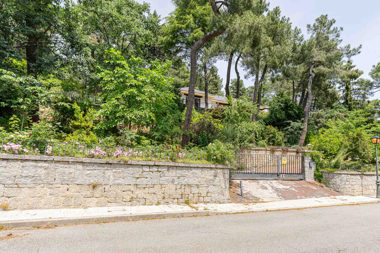 Exterior view of a house on a hill, with a stone wall, metal gate, and lush green trees.