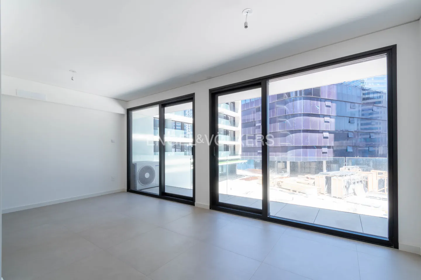 Bright, empty room with white walls, gray tile floor, and large black-framed sliding glass doors overlooking a modern building.