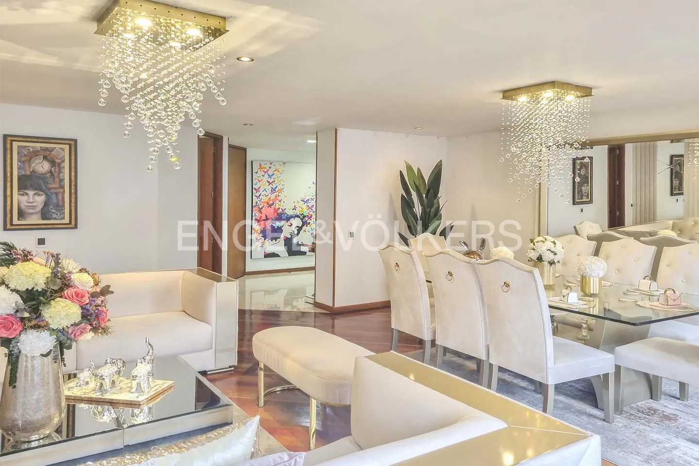 Living room with white sofas, a glass table, and a dining area with beige chairs and a glass table. Crystal chandeliers hang from the ceiling.