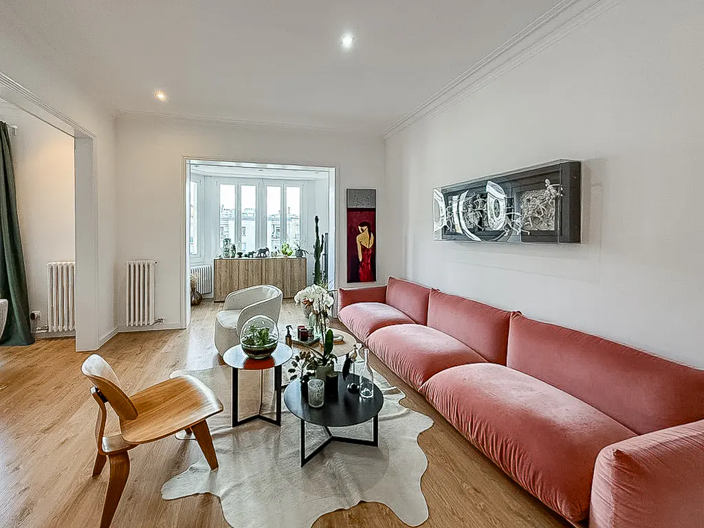 Modern living room with a large pink velvet sofa, wooden chair, two round coffee tables, and a cowhide rug on wood floors.