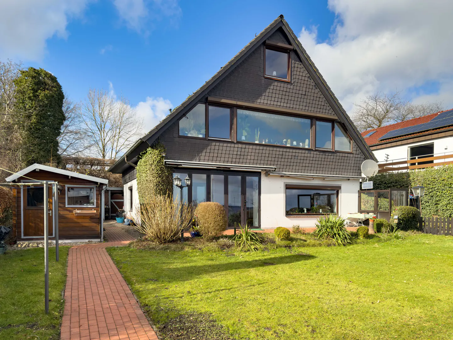 A two-story house with a dark roof and white siding, surrounded by a green lawn and a brick path leading to a wooden shed.