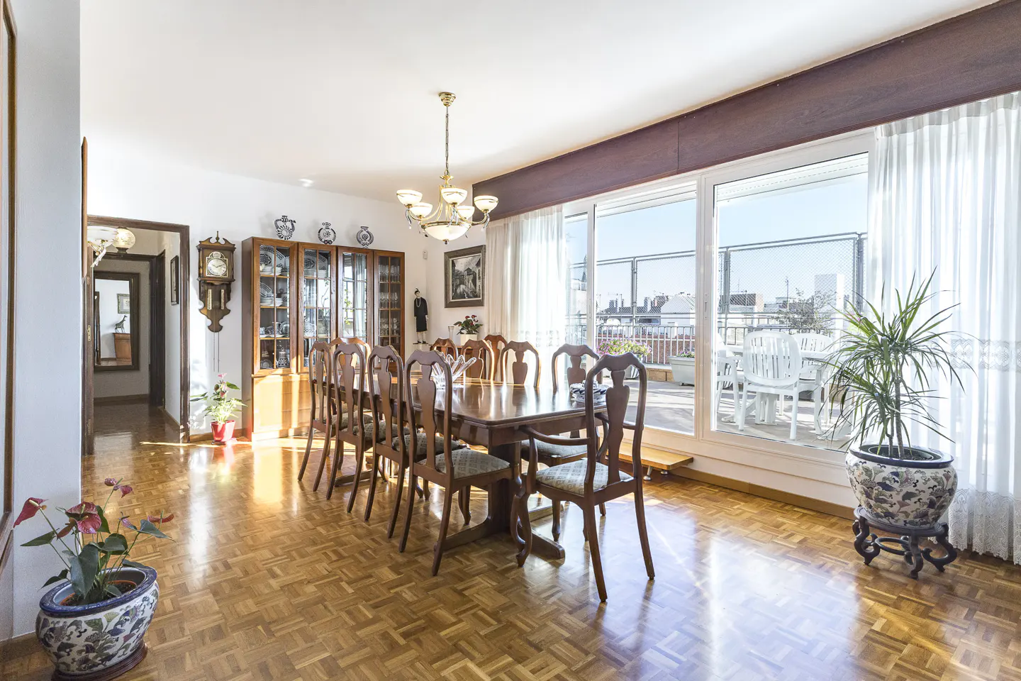 Bright dining room with a long wooden table, chairs, chandelier, and a view of a balcony with white furniture.