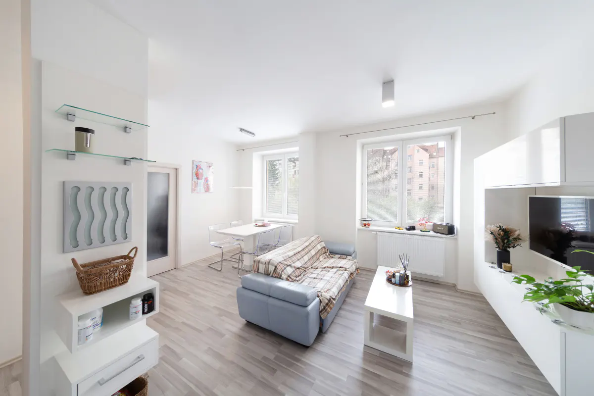 Bright, modern living room with a blue sofa, white furniture, and light wood floors. Windows offer natural light.