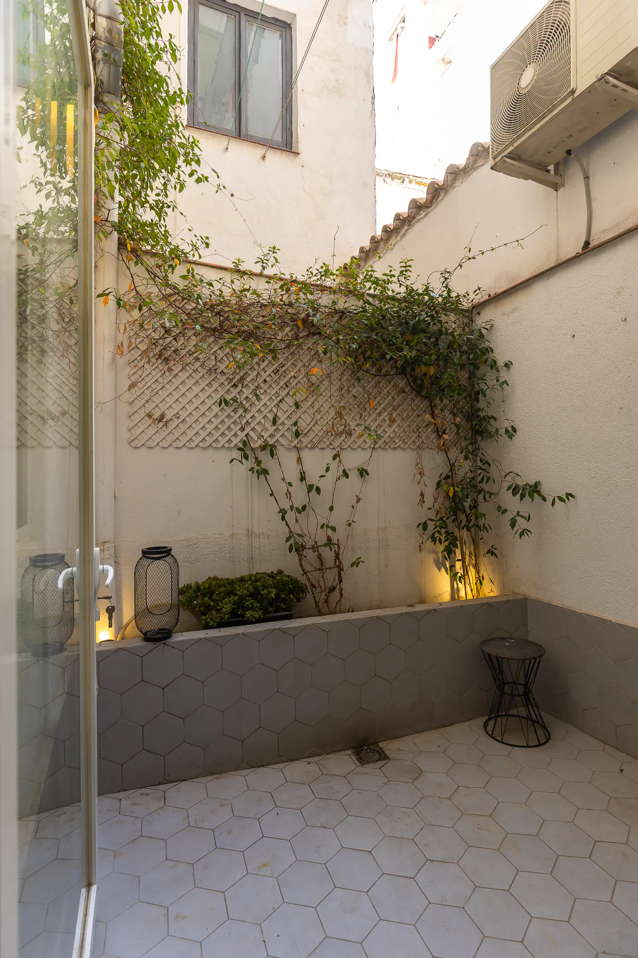 A small courtyard with white walls, climbing plants, and gray hexagonal tiles. A black metal table sits in the corner.