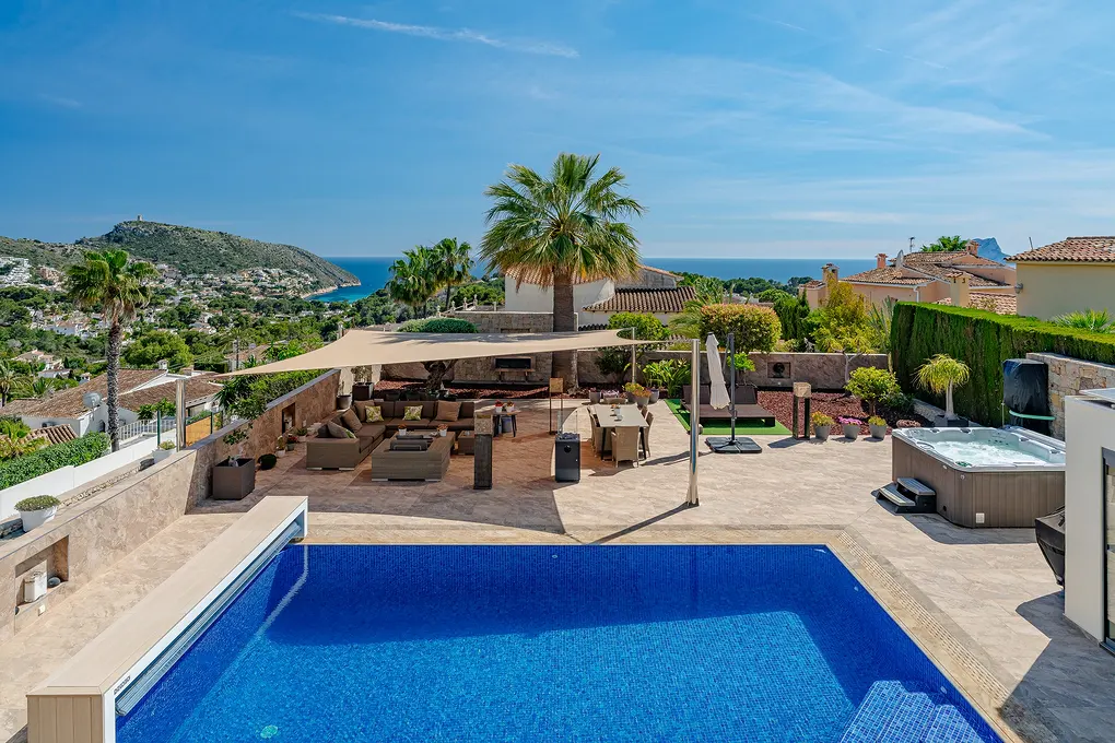 Luxury villa with a blue tiled pool, outdoor seating, a hot tub, and a view of the ocean and mountains under a clear blue sky.