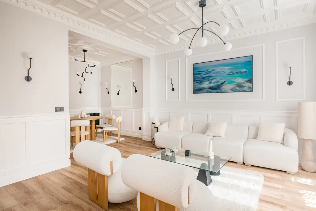 Bright, modern living room with white walls, wood floors, and white furniture. A blue abstract painting hangs above a white sofa.