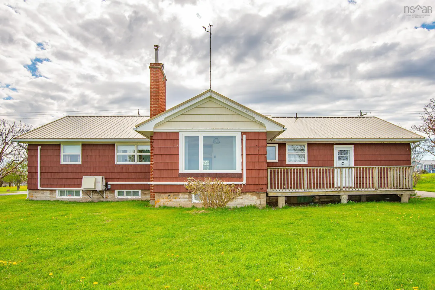 Red bungalow with a metal roof, brick chimney, and wooden deck, set against a cloudy sky over a green grassy lawn.