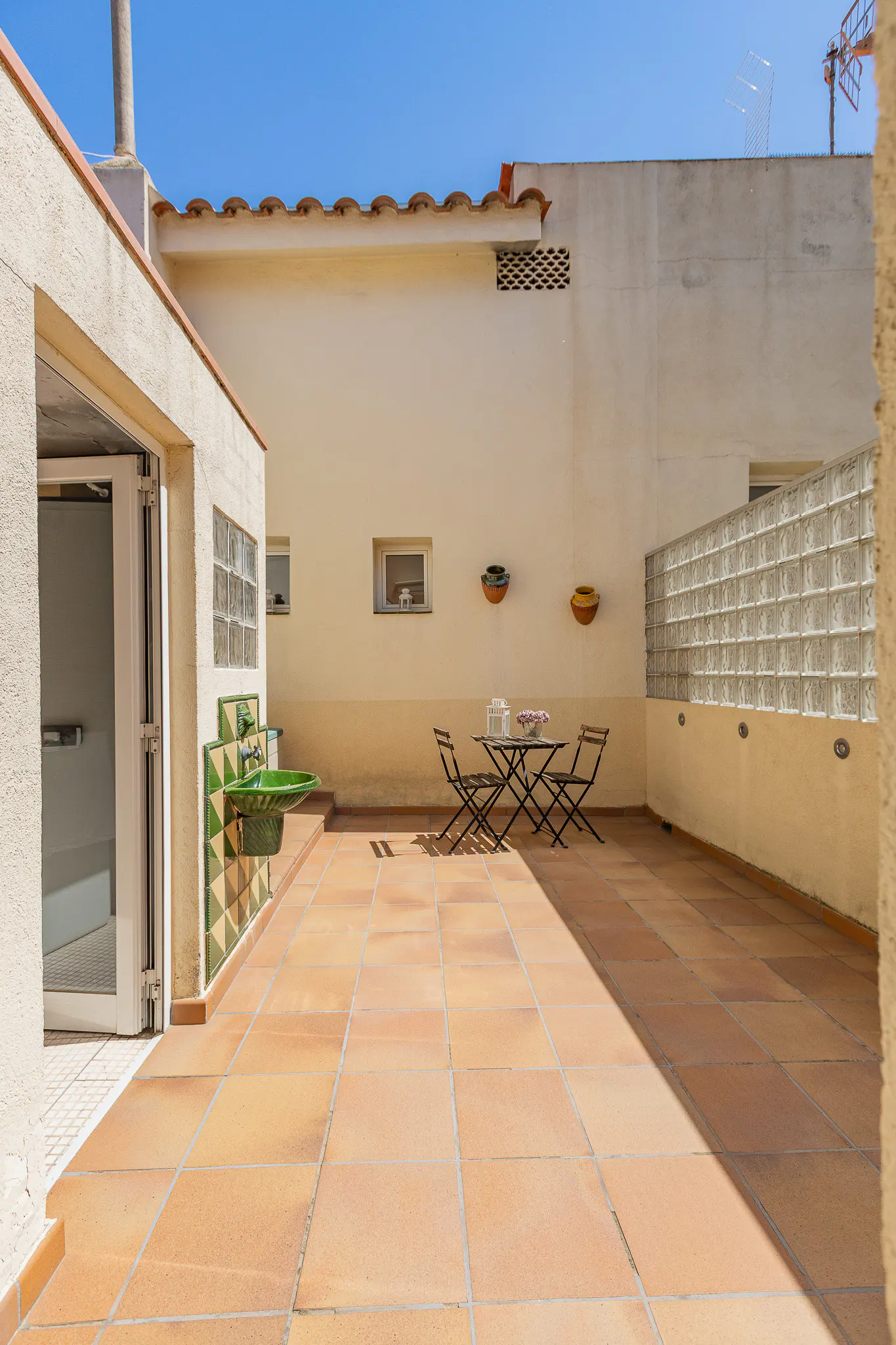 Outdoor patio with terracotta tiles, a table and chairs, a green sink, and a glass block wall under a clear blue sky.