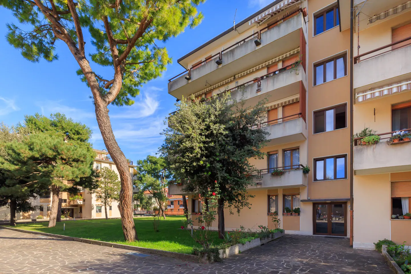 Exterior view of a tan apartment building with balconies, green trees, and a stone driveway under a blue sky.
