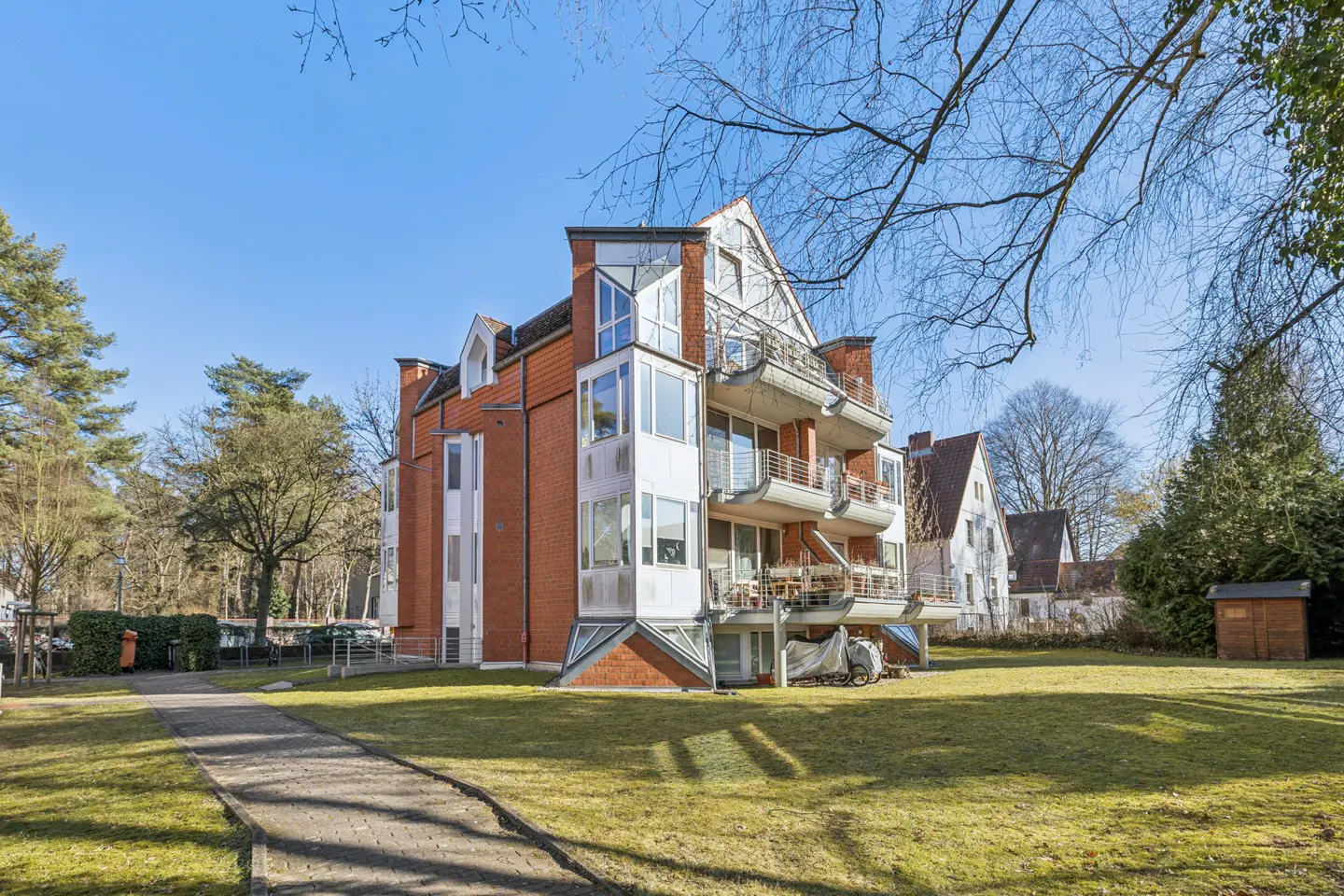 Exterior view of a red brick apartment building with balconies, surrounded by a green lawn and trees under a blue sky.