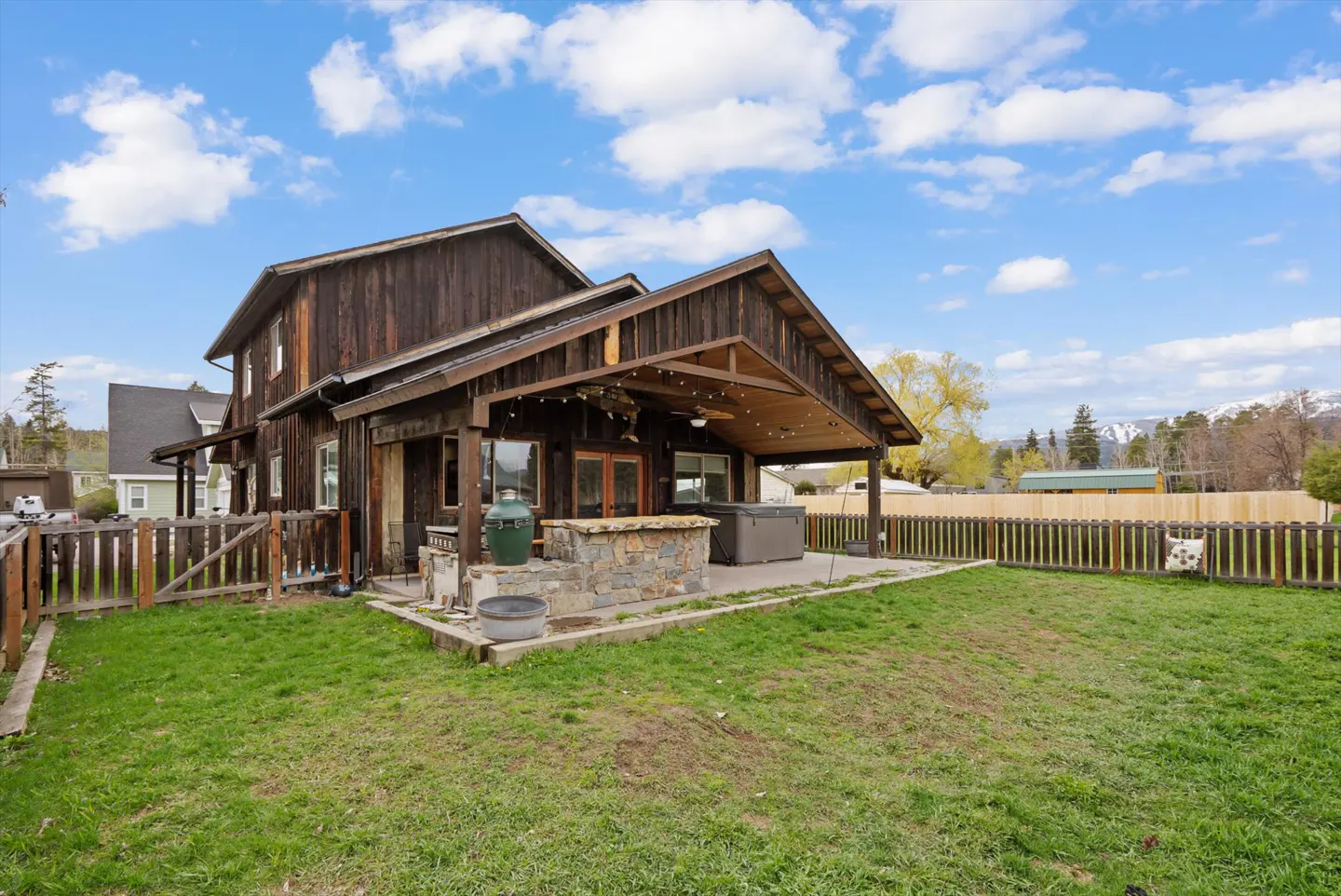 Exterior view of a two-story brown house with a patio, grill, hot tub, and green lawn under a blue sky.