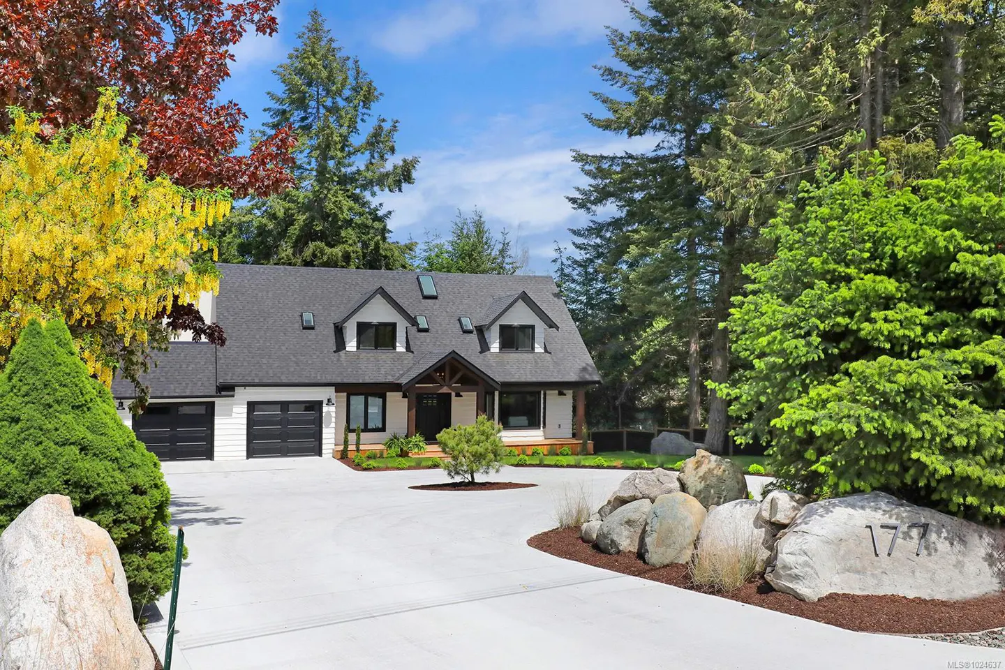 A modern white house with a black roof and garage doors, surrounded by lush greenery and a concrete driveway.