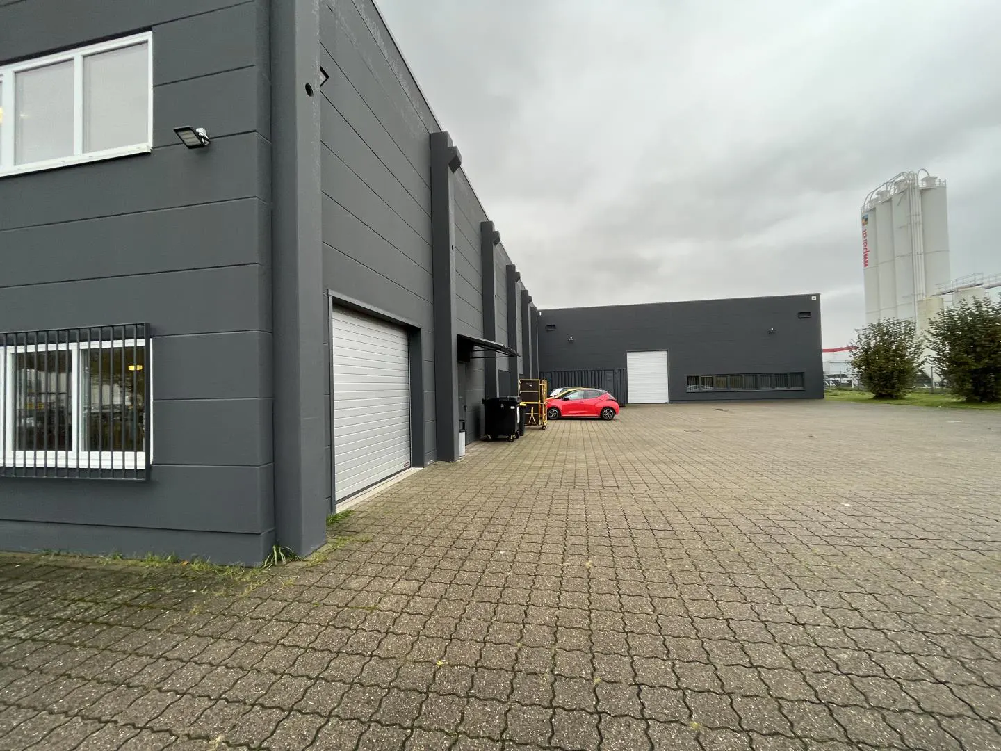 Exterior of a gray industrial building with a closed garage door and a red car parked in the paved lot.
