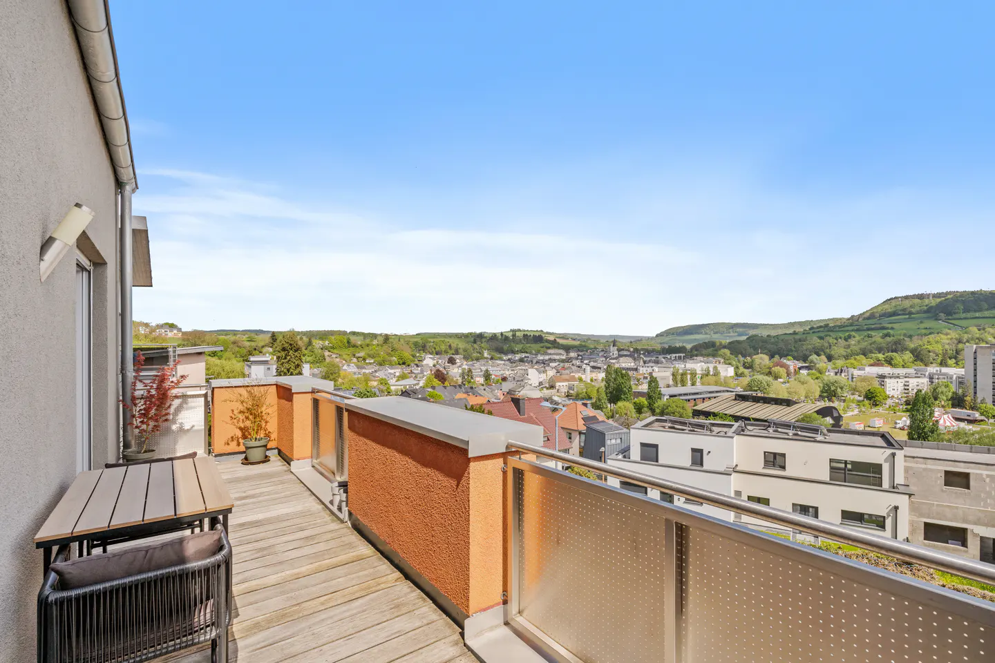 Balcony view with table, chair, and orange wall overlooking a town and green hills under a blue sky.