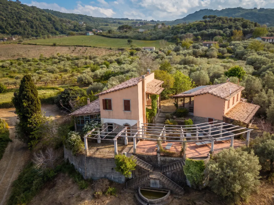Aerial view of a peach-colored villa with a terracotta roof, surrounded by olive trees and green hills. A pergola covers a patio with stairs leading to a fountain.