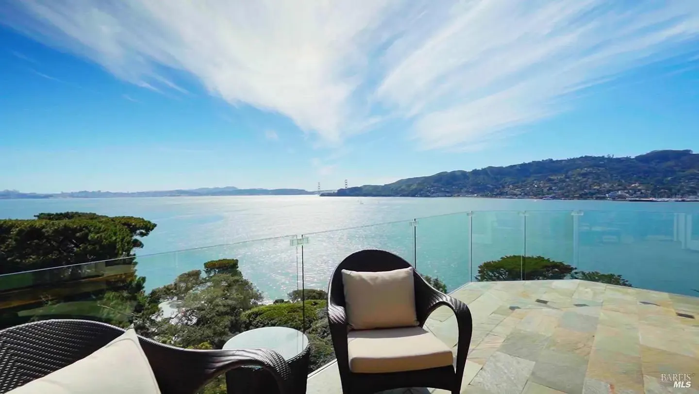 Outdoor patio with wicker furniture and a glass railing overlooking the San Francisco Bay and the Golden Gate Bridge on a sunny day.