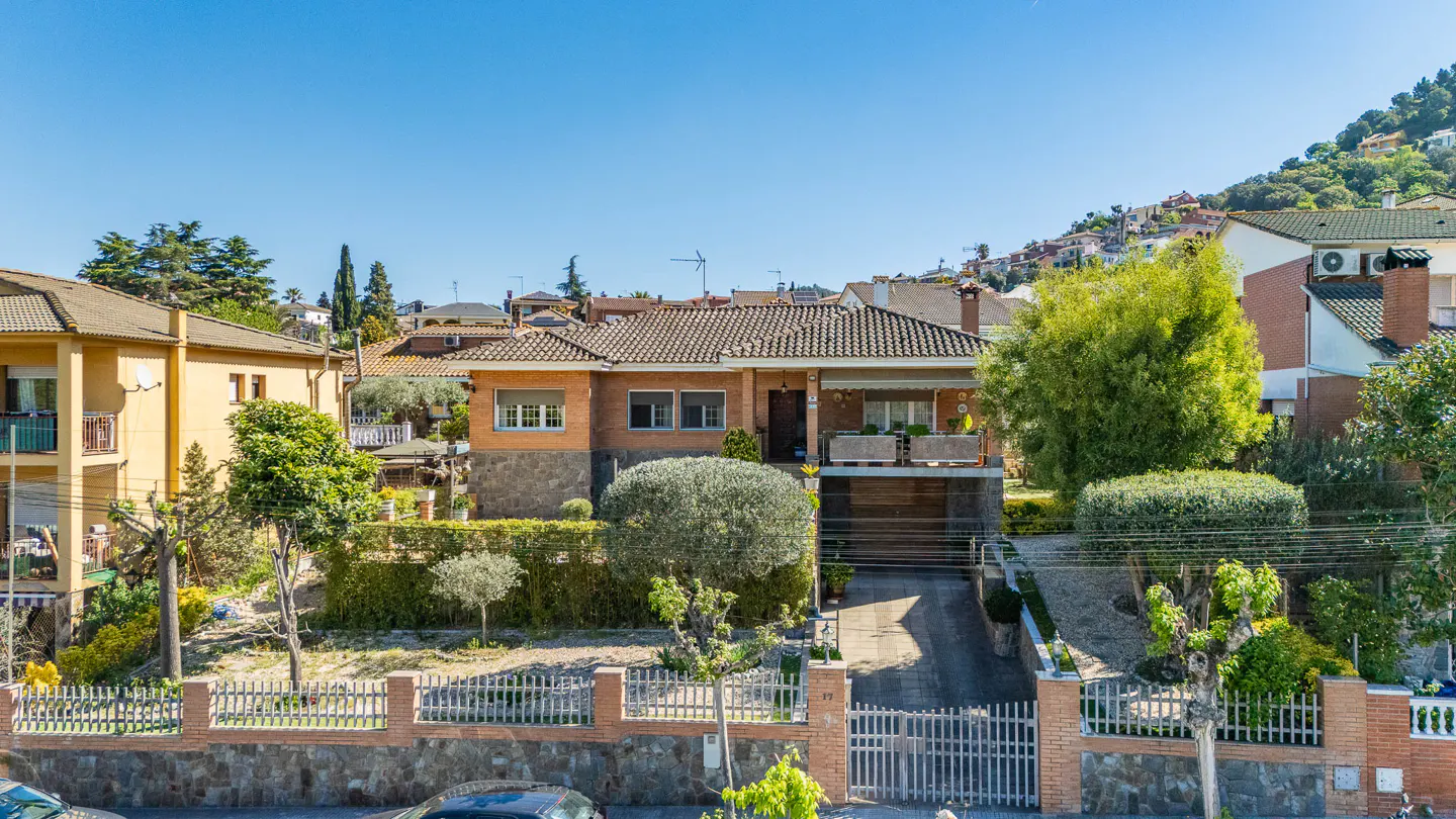 Exterior view of a two-story brick house with a tile roof, a gated driveway, and lush green landscaping.