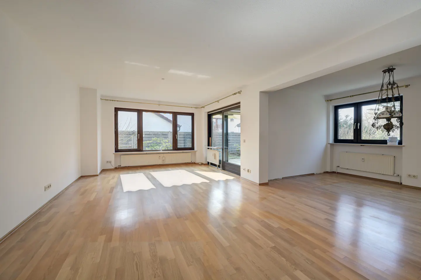 Bright, empty living room with hardwood floors, white walls, and large windows. A chandelier hangs near a window with a radiator below.