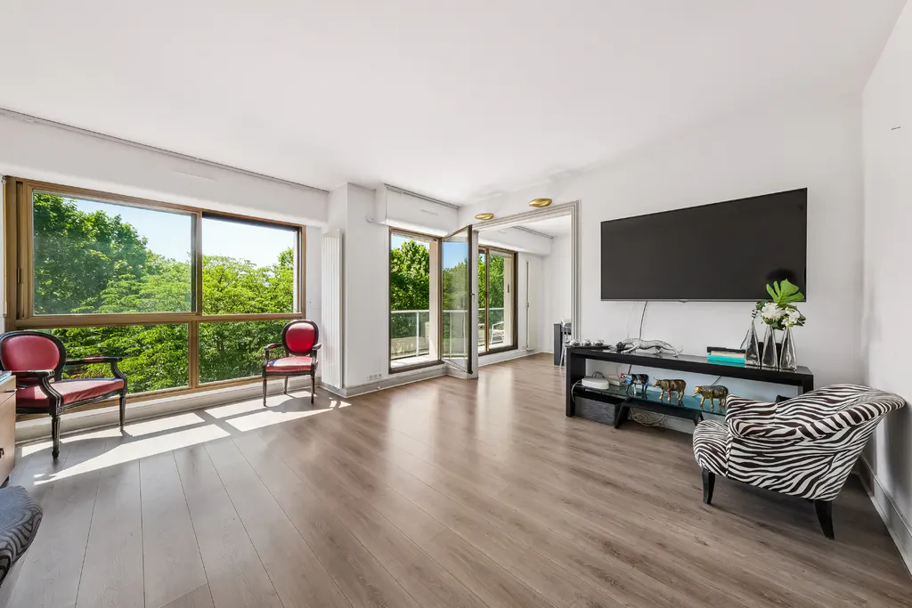 Bright living room with wood floors, large windows overlooking trees, red chairs, TV, and zebra print chair.