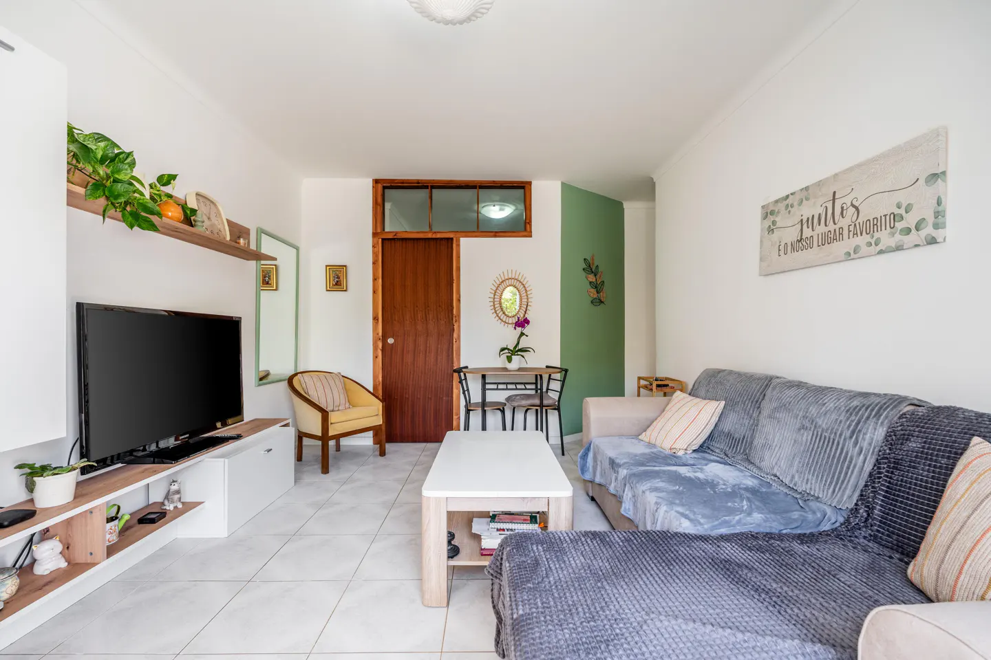A bright living room with a gray sofa, white coffee table, TV, and dining area. The walls are white with green accents.